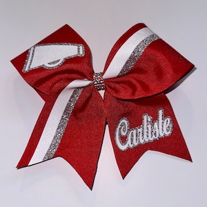 May include: Red cheer bow with white and silver glitter stripes. The bow has a white megaphone graphic and the name "Carliste" in silver glitter.