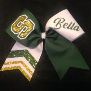 May include: Green and white cheer bow with gold glitter accents. The bow has a green section with a gold glitter "S" and a white section with the name "Bella" in gold glitter.