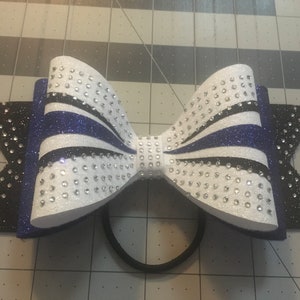 May include: A white, black, and blue cheer bow with rhinestones. The bow is tied with a black elastic band.