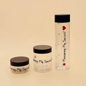 May include: Three clear plastic containers with black lids of varying heights. Each container features the black text "Measuring My Success!" and the tallest bottle includes a red heart and star icon with vertical measurement markings in millilitres.