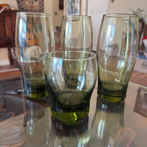 Peut inclure: Quatre verres à boire teintés de vert de différentes tailles. Deux sont grands et fins, un est court et arrondi, et un autre est de taille moyenne. Les verres sont disposés sur une surface réfléchissante, mettant en valeur leur design transparent.