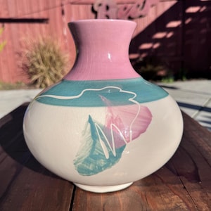 Puede incluir: Un jarrón de cerámica con cuerpo bulboso y cuello estrecho. El jarrón presenta una combinación de colores rosa, turquesa y blanco roto. El cuerpo tiene un diseño floral en rosa y turquesa. El jarrón está sobre una superficie de madera.