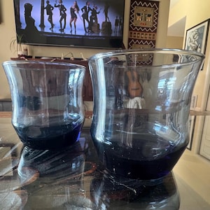Puede incluir: Dos vasos de vidrio teñido de azul de diferentes tamaños. Los vasos tienen forma acampanada y una base azul oscuro. Los vasos están sobre una superficie reflectante, el más grande a la derecha y el más pequeño a la izquierda.