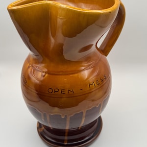 Könnte beinhalten: Vintage Keramikkrug mit einer einzigartigen Form und glänzender, brauner Glasur. Der Krug hat einen breiten, wellenförmigen Rand und einen Henkel. Die Worte "OPEN - MESS" sind auf dem Körper eingraviert. Der Sockel ist in einem dunkleren Braunton gehalten.
