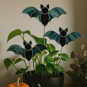 May include: Three decorative bat plant stakes with teal and black stained glass wings are displayed in a black pot with a green plant. Two pumpkins and a brass bell are on a wooden surface. Halloween decor.