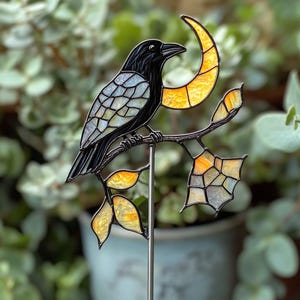 Puede incluir: Un cuervo de vidriera posado en una rama con una luna creciente. El cuervo es negro con alas iridiscentes, y la luna y las hojas son amarillas y naranjas. La pieza está montada en una estaca de metal para jardín.