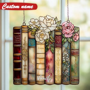 May include: Stained glass panel featuring a stack of books with white and pink roses. The books have ornate gold detailing and the name "Emily" is visible. The panel is suspended by a chain and has the text "Custom name" at the top.
