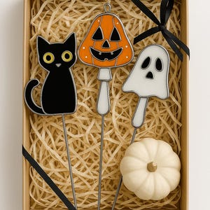 May include: A decorative Halloween-themed gift set in a cardboard box with straw filler. The set includes a black cat, jack-o'-lantern, and ghost garden stakes, along with a small white pumpkin. A black ribbon decorates the box.
