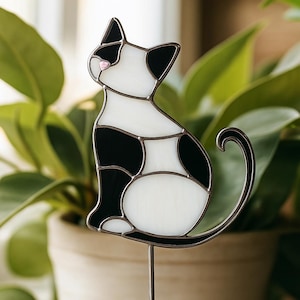 Può includere: Un segnaposto da giardino a forma di gatto in vetro colorato bianco e nero. Il gatto è seduto con la coda arricciata. Ha un naso rosa ed è montato su un paletto di metallo. Lo sfondo è una pianta verde sfocata.