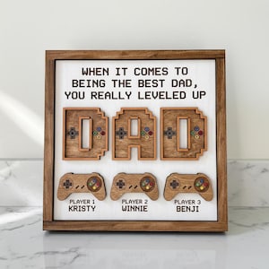 May include: Wooden framed sign with the word "DAD" in pixelated letters, each with a game controller design. Below are three controller designs with the names "Kristy", "Winnie", and "Benji". The sign reads "When it comes to being the best dad, you really leveled up."
