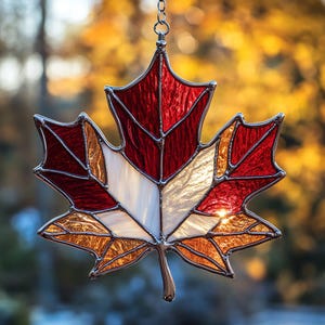 May include: Stained glass maple leaf suncatcher with red, white, and gold sections, framed with a silver metal border. The leaf is suspended by a chain, showcasing a fall theme.