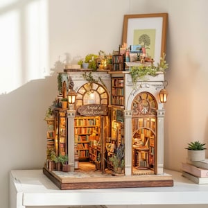 Op de afbeelding: Een miniatuurmodel van een boekwinkel met de tekst "Anna's Bookstore" boven de ingang. Het model heeft gedetailleerde boekenplanken, gewelfde ramen en kleine hangende lantaarns. Groen en kleine planten sieren de buitenkant, met een warme gloed van binnenuit.