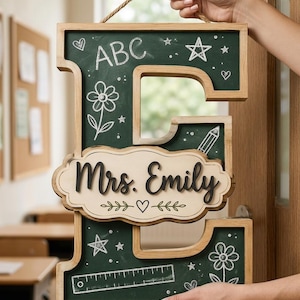 May include: A wooden letter E sign with a dark green chalkboard finish. The surface features white chalk-style drawings of flowers, stars, and a ruler. A central cream-colored plaque displays the black text Mrs. Emily with a heart icon.