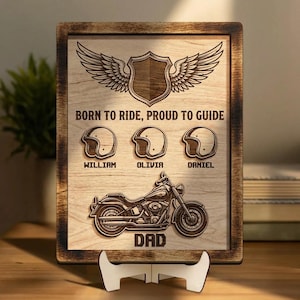 May include: Wooden plaque with a motorcycle design, helmets, and the text "BORN TO RIDE, PROUD TO GUIDE." The plaque includes the names William, Olivia, and Daniel, and the word "DAD." The plaque is displayed on a white stand.