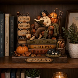 Peut inclure: Une figurine décorative représentant une personne lisant dans un fauteuil en cuir sur une pile de livres étiquetés "JUST A GIRL WHO LOVES BOOKS". La base porte l'inscription "Sophia's Reading Nook" avec une citrouille, un chat et une lampe.