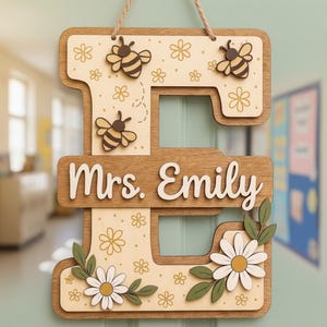 Könnte beinhalten: Holzschild in Form des Buchstabens „E“ mit dem Namen „Mrs. Emily“. Das Schild ist mit Bienen, Gänseblümchen und floralen Akzenten verziert. Es hängt an einem Naturseil.
