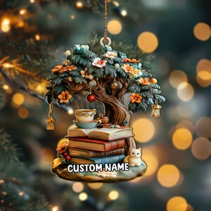 May include: A decorative ornament featuring a tree with orange and white flowers, a stack of books, a teacup, and a small white cat. The ornament has a gold hanging loop and the text "CUSTOM NAME" at the bottom.