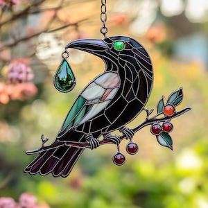 Könnte beinhalten: Glasfenster-Suncatcher mit einer schwarzen Krähe, einem grünen Auge und einem grünen Tropfen im Schnabel. Die Krähe sitzt auf einem Ast mit roten Beeren und grünen Blättern. Der Suncatcher hängt an einer Metallkette.