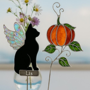 Peut inclure: Silhouette d'un chat noir avec des ailes de fée irisées et une petite plaque nominative « Lis » à côté d'une décoration de citrouille en vitrail. La citrouille est orange et rouge avec des feuilles vertes, parfaite pour l'automne ou Halloween.