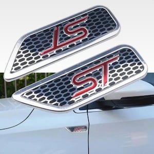 Op de afbeelding: Twee zilveren en zwarte auto-emblemen met de letters "ST" in rood. De emblemen hebben een honingraatpatroon en zijn bevestigd op een witte auto. De zijspiegel van de auto is zichtbaar.