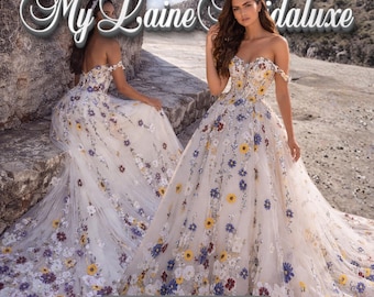 Puede incluir: Dos vestidos de novia blancos con bordados florales. Los vestidos presentan escotes sin hombros y están adornados con flores de colores. La imagen incluye el texto "My Laine Bridaluxe" y "Designed for Your Happily Ever After."