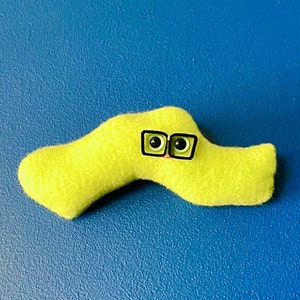 May include: A lime green felt creature with black-rimmed glasses and black eyes. The plush toy is set against a blue background. The creature has a curved, worm-like shape.