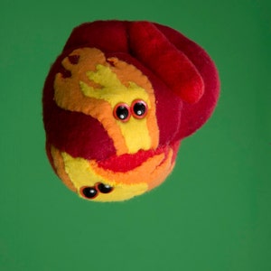 Puede incluir: Una criatura de peluche caprichosa con un cuerpo rojo y acentos amarillos, naranjas y rojos. Tiene dos juegos de ojos negros y una boca sonriente. El juguete est&aacute; sobre un fondo verde liso.