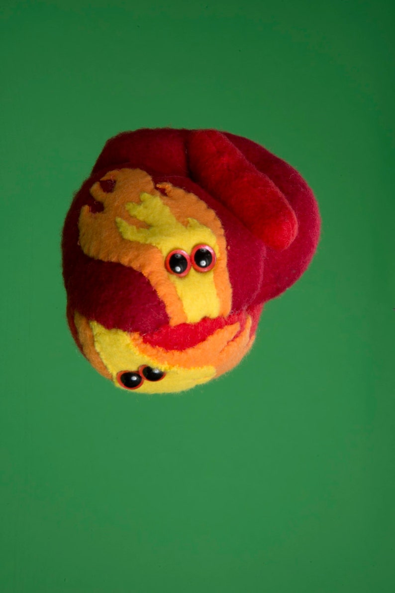 Puede incluir: Una criatura de peluche caprichosa con un cuerpo rojo y acentos amarillos, naranjas y rojos. Tiene dos juegos de ojos negros y una boca sonriente. El juguete est&aacute; sobre un fondo verde liso.