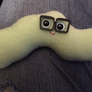 May include: A light green plush worm-like creature with large, square-framed glasses and a stitched red smile. The toy has black eyes and is made of a soft, textured material. The creature is a whimsical, handmade item.