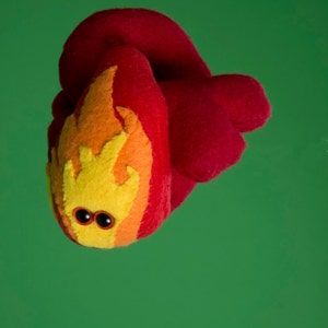 Puede incluir: Una criatura de fieltro hecha a mano con forma de llama, con un cuerpo rojo y un dise&ntilde;o de llama amarillo, naranja y rojo. Tiene dos peque&ntilde;os ojos saltones negros. El fondo es verde liso.