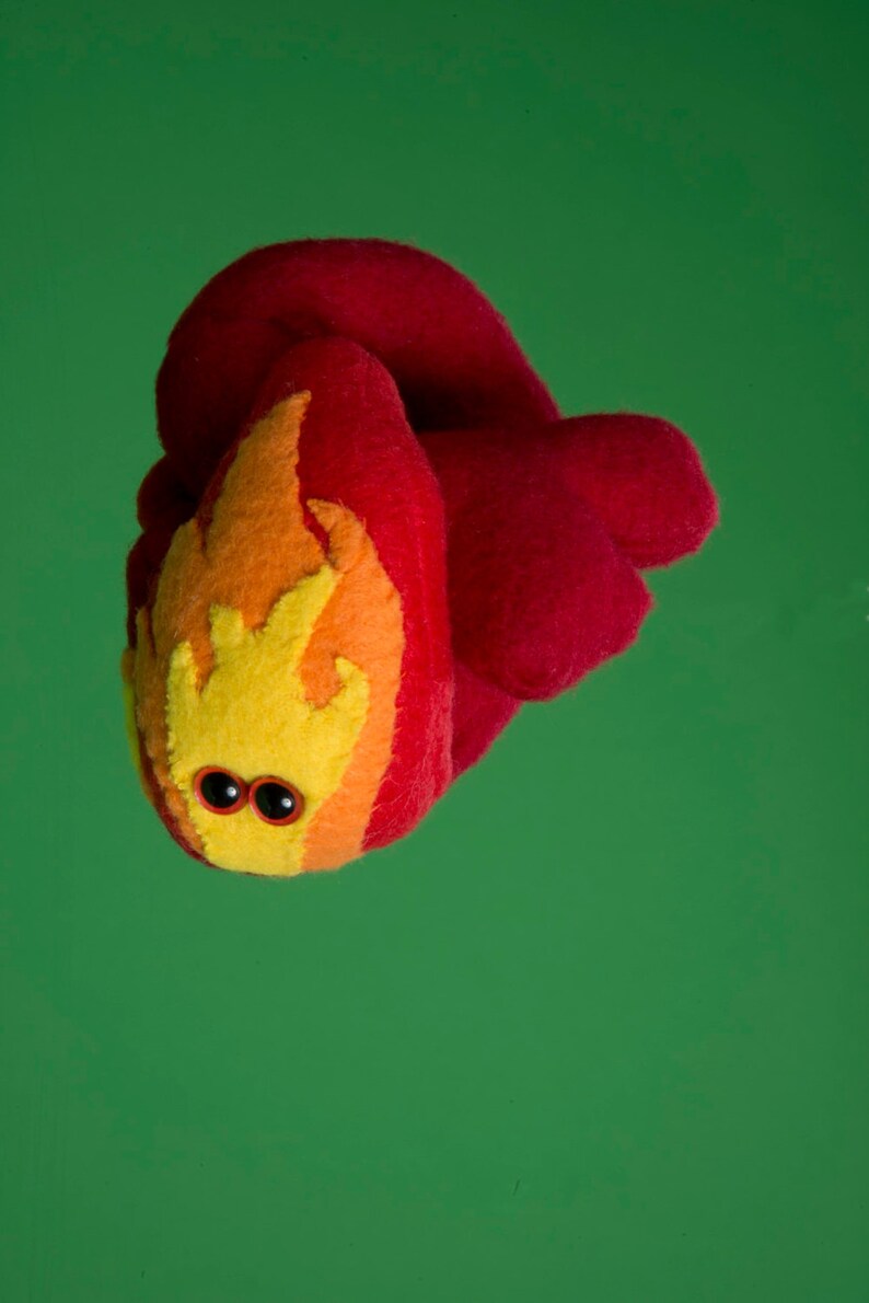 Puede incluir: Una criatura de fieltro hecha a mano con forma de llama, con un cuerpo rojo y un dise&ntilde;o de llama amarillo, naranja y rojo. Tiene dos peque&ntilde;os ojos saltones negros. El fondo es verde liso.