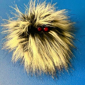 Könnte beinhalten: Ein handgefertigtes, flauschiges Wesen mit einer Mischung aus schwarzem, weißem und hellbraunem Fell. Es hat zwei kleine, runde, rote Augen. Das Wesen ist vor einem einfarbigen blauen Hintergrund platziert.
