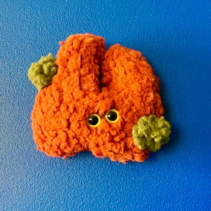 May include: A handmade orange plush toy with two googly eyes and two small green pom-poms. The toy has a textured, fuzzy appearance against a blue background.