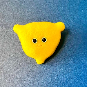 Puede incluir: Un juguete de peluche con forma de limón amarillo brillante, con ojos negros y blancos y una pequeña sonrisa. El peluche tiene dos pequeñas protuberancias en forma de oreja en la parte superior. El juguete está sobre un fondo azul texturizado.