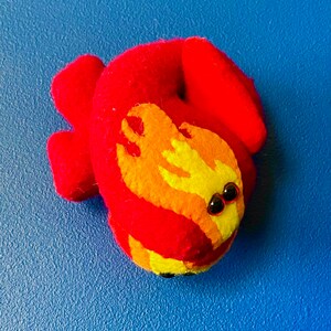 Puede incluir: Una criatura de fieltro hecha a mano en tonos rojos, naranjas y amarillos, con peque&ntilde;os ojos de cuentas negras. El juguete de fieltro est&aacute; sobre una superficie azul. El dise&ntilde;o se asemeja a un drag&oacute;n o salamandra estilizada.
