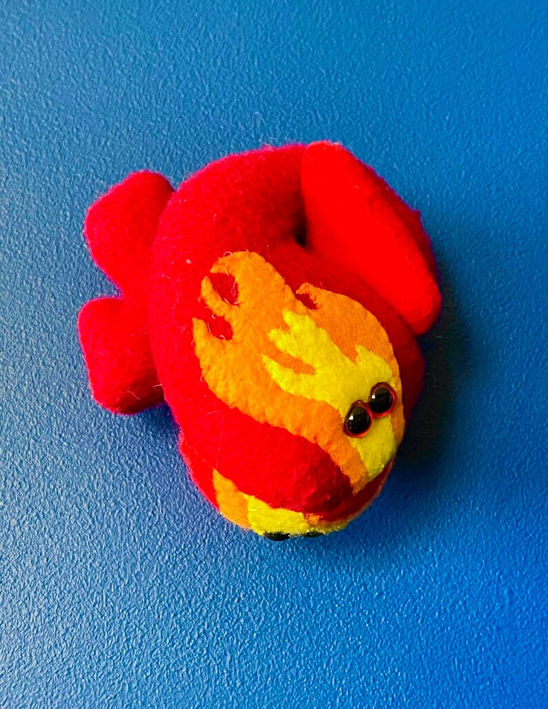 Puede incluir: Una criatura de fieltro hecha a mano en tonos rojos, naranjas y amarillos, con peque&ntilde;os ojos de cuentas negras. El juguete de fieltro est&aacute; sobre una superficie azul. El dise&ntilde;o se asemeja a un drag&oacute;n o salamandra estilizada.