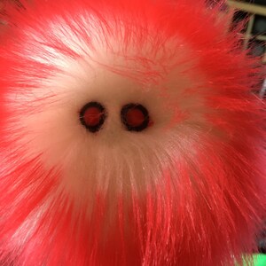 May include: Close-up of a fuzzy, plush toy with bright red fur and a white face. The toy has two small, round, red eyes. The fur is long and shaggy, giving the toy a playful appearance. The toy is a vibrant red and white color.