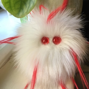 May include: A whimsical, handmade creature with a fluffy white body and bright red accents. Two red, round eyes give it a playful look. The creature has red strands of material extending from its body, adding to its unique design.