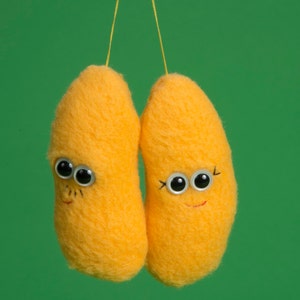 May include: Two yellow, bean-shaped plush toys with googly eyes, eyelashes, and smiling faces hang against a green backdrop. They are suspended by thin yellow strings, creating a whimsical and playful aesthetic.