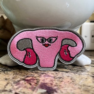 May include: Embroidered patch featuring a stylized pink uterus with a face, gray accents, and a silver border. The patch has a cartoonish design with a red heart-shaped mouth and black eyes. This patch is suitable for clothing or accessories.