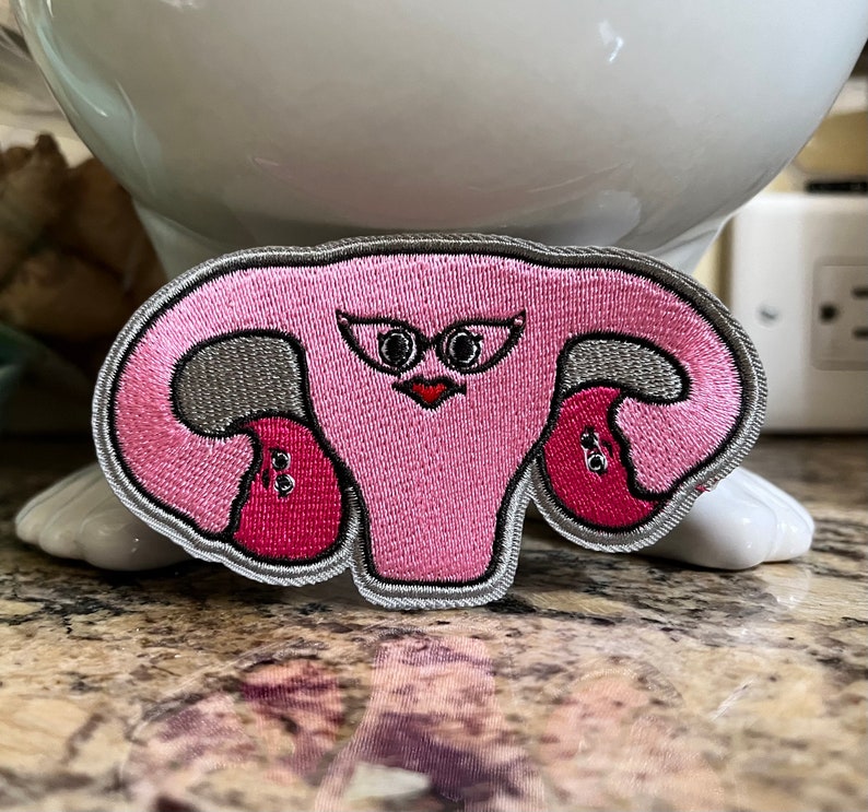 May include: Embroidered patch featuring a stylized pink uterus with a face, gray accents, and a silver border. The patch has a cartoonish design with a red heart-shaped mouth and black eyes. This patch is suitable for clothing or accessories.