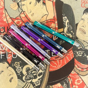 Könnte beinhalten: Eine Auswahl an farbenfrohen Stiften in verschiedenen Farbtönen, darunter Türkis, Blau, Lila, Pink, Schwarz und Creme. Die Stifte sind vor dem Hintergrund eines japanischen Vintage-Holzschnitts angeordnet. Jeder Stift hat einen silberfarbenen Clip und eine Spitze.