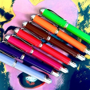 Könnte beinhalten: Ein Set aus sieben Füllfederhaltern mit bunten Gehäusen. Die Stifte sind in einem Regenbogenmuster angeordnet, wobei der grüne Stift oben und der violette Stift unten ist. Die Stifte haben silberne Akzente und sind aus einem glatten, glänzenden Material gefertigt.