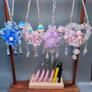May include: A display of handmade beaded necklaces with floral and character pendants. The necklaces feature various colors, including blue, pink, and purple, with dangling crystal accents. The necklaces are hanging on a wooden display stand.