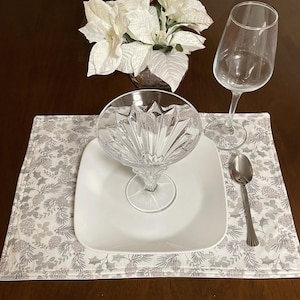 May include: A festive table setting featuring a white square plate, a crystal dessert dish, a wine glass, and a spoon. The place setting rests on a placemat with a gray and white botanical pattern. A vase of white poinsettias adds a seasonal touch.
