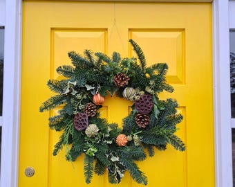 May include: A festive Christmas wreath hangs on a bright yellow door. The wreath is made of evergreen branches, pinecones, and dried orange slices. The door has a black mail slot and a white trim around the door frame.