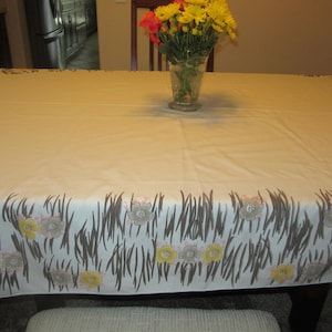 May include: A rectangular tablecloth with a cream-colored base featuring a decorative border pattern of dark brown vertical grass-like lines interspersed with yellow and light gray floral motifs along the edges of the fabric.
