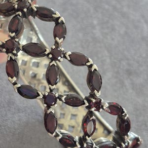 Op de afbeelding: Zilverkleurige armband met een herhalend bloemmotief met ovaal geslepen, dieprode edelstenen. De armband heeft een filigraan patroon. De edelstenen zijn in een symmetrisch patroon gerangschikt.