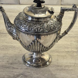 Op de afbeelding: Een zilveren theepot met een sierlijke, in reliëf aangebrachte bloemenband rond de bovenkant en het deksel. De voet en het onderste deel van de romp zijn versierd met verticale groeven. De pot heeft een gebogen tuit, een lichtgekleurd handvat en een zwarte knop.