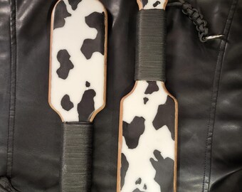 May include: Two paddles with a cow print design. The paddles have a white base with black spots, a wooden border, and a black wrapped handle. Each paddle has a black lanyard attached to the handle.